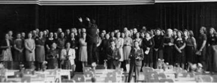 Black and white photo shows a group of about 50 people in a choir with conductor in front of them.
