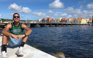 Young man in sunglasses sitting on a pier next to water. A bridge and buildings are on the opposite side of the water in the background.