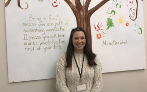 Young woman standing in front of a piece of artwork including text, a tree, and hand prints. Text reads, "Being a family means you are part of something wonderful. It means you will love and be loved for the rest of your life. No matter what."