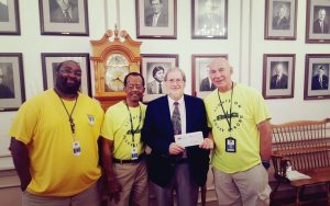 Three men in yellow shirts standing with an older man in a suit holding a check