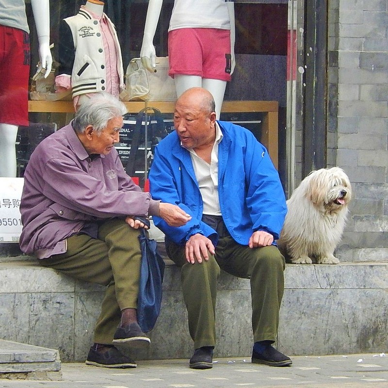 Two older men sit together in front of a shop having a conversation.