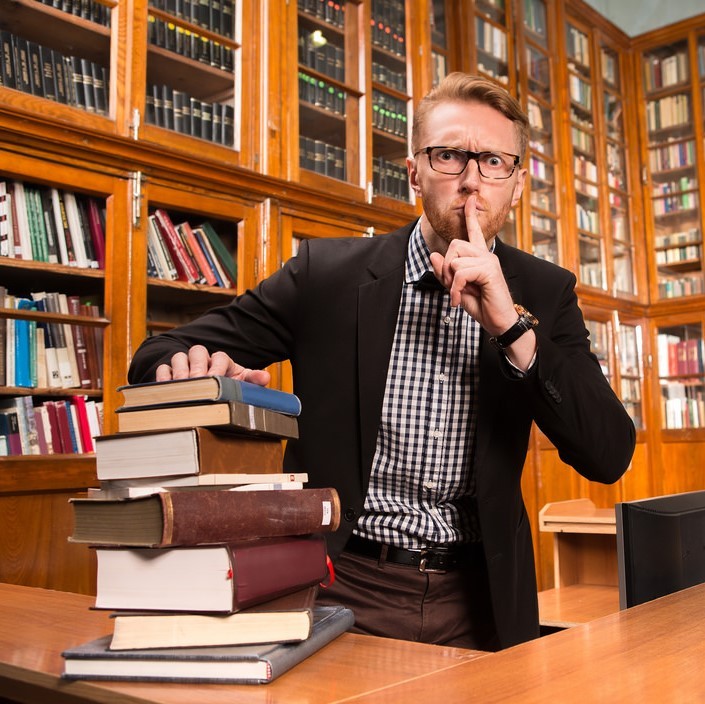 A man dressed in a jacket and bow tie stands behind a desk stacked with books. He has a stern look on his face and is putting a finger to his lips requesting quiet. Behind the man are shelves lined with books.