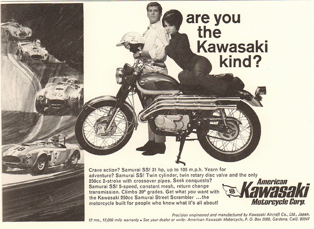 An advertisement for Kawasaki motorcycles from the 1970's. The slogan is "Are You the Kawasaki Kind?" The ad features an average looking man dressed in a shirt and tie standing behind a Kawasaki motorcycle and holding a motorcycle helmet. An attractive woman sits on the seat of the motorcycle and leans her head on the man's shoulder.