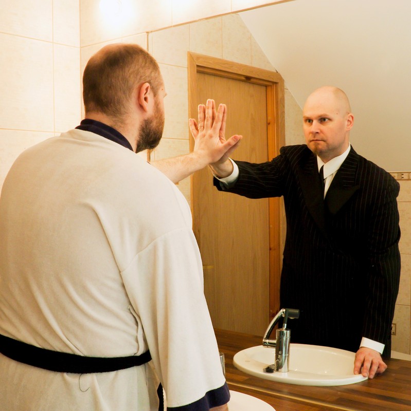 A man stands in front of the bathroom mirror and reaches out to touch an altered reflection of himself.