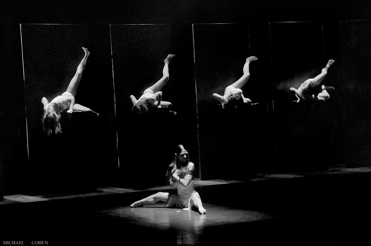 A dancer sits on stage in a spotlight with 4 dancers in the background laying on 3-foot-tall boxes.