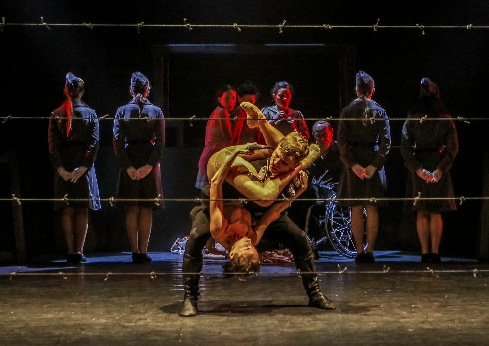 Photo of dancers on stage behind barbed wire fencing