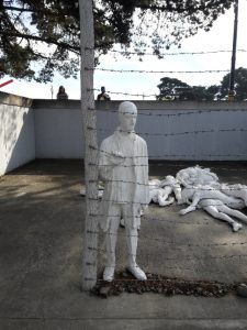 Different angle of the same sculpture of man standing at barbed-wire fence near a pile of sculpted bodies.