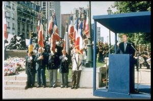 Color photo of Chirac at a podium with men next to him holding flags.