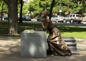 A bronze statue of Phillis Wheatley, a young African-American woman. The statue is placed with a stone cube with information about Wheatley carved into it. She leans against it, making the folds of her dress pronounced. She rests her head on her left hand, contemplating.