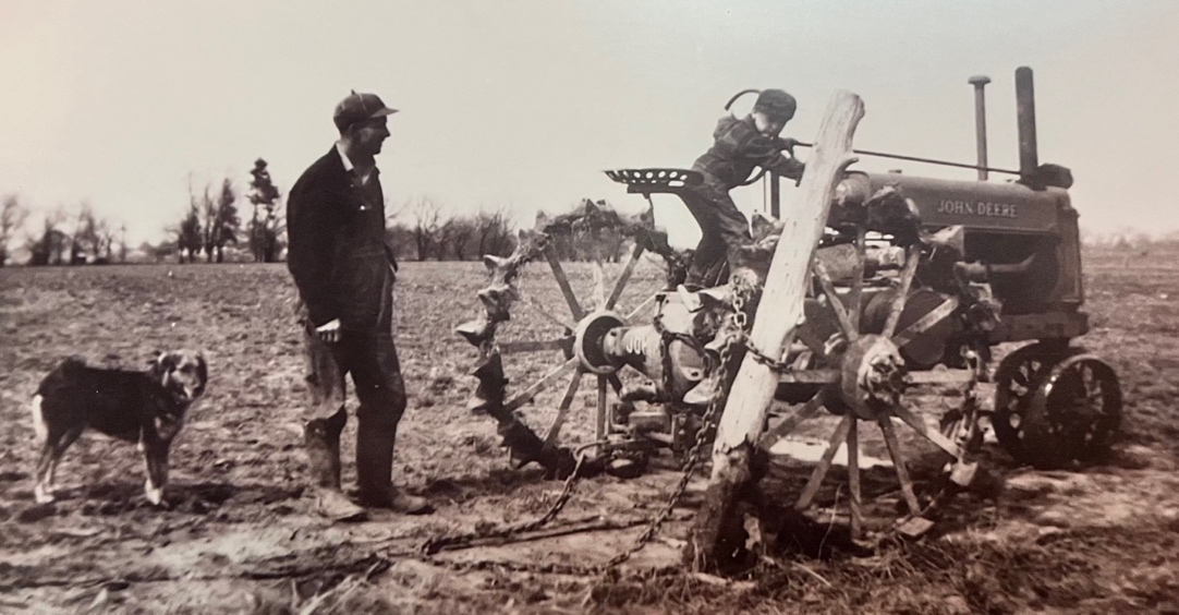 Man and child with a vintage tractor and a dog in a field.