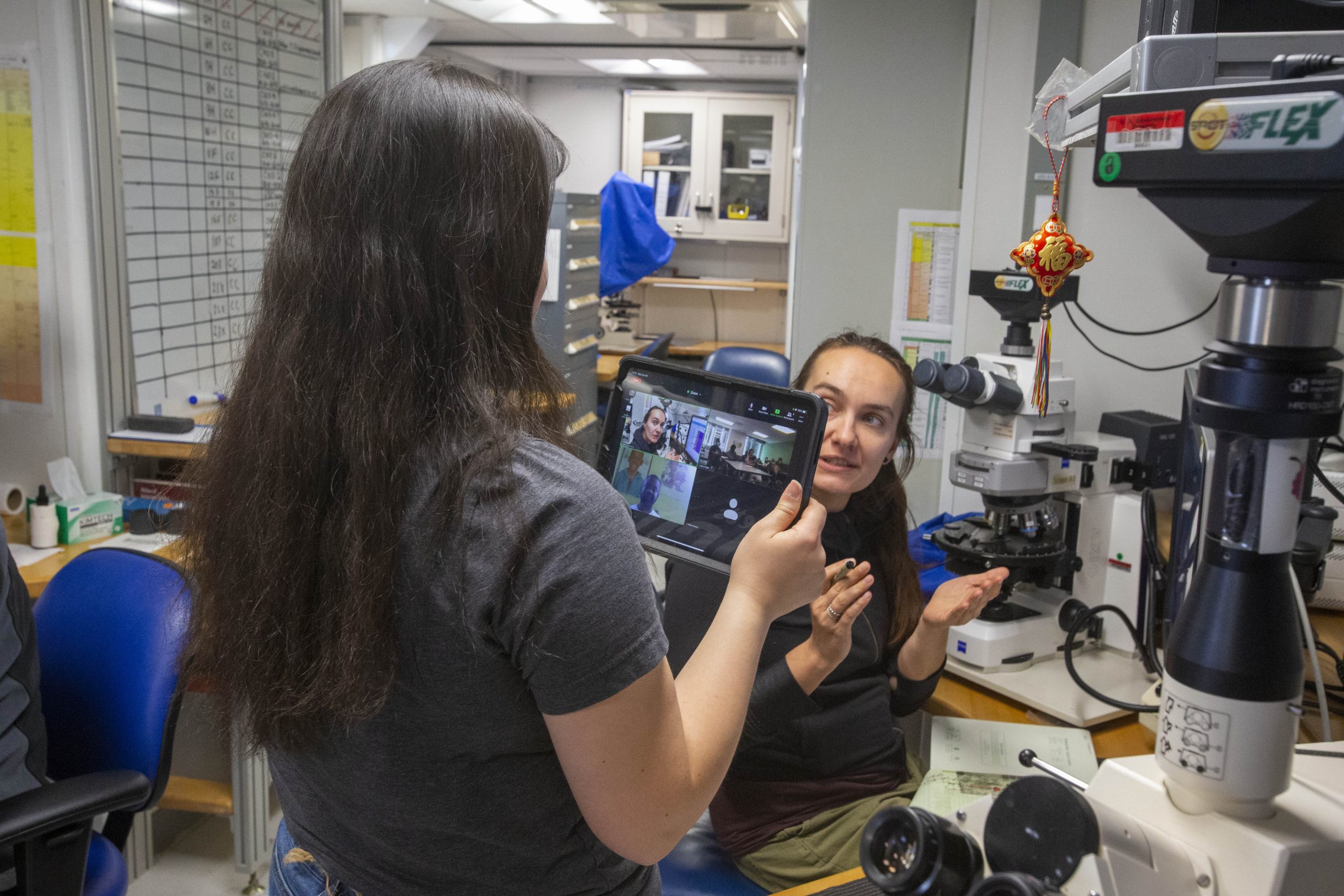 Person holding an iPad talking to another person sitting at a microscope