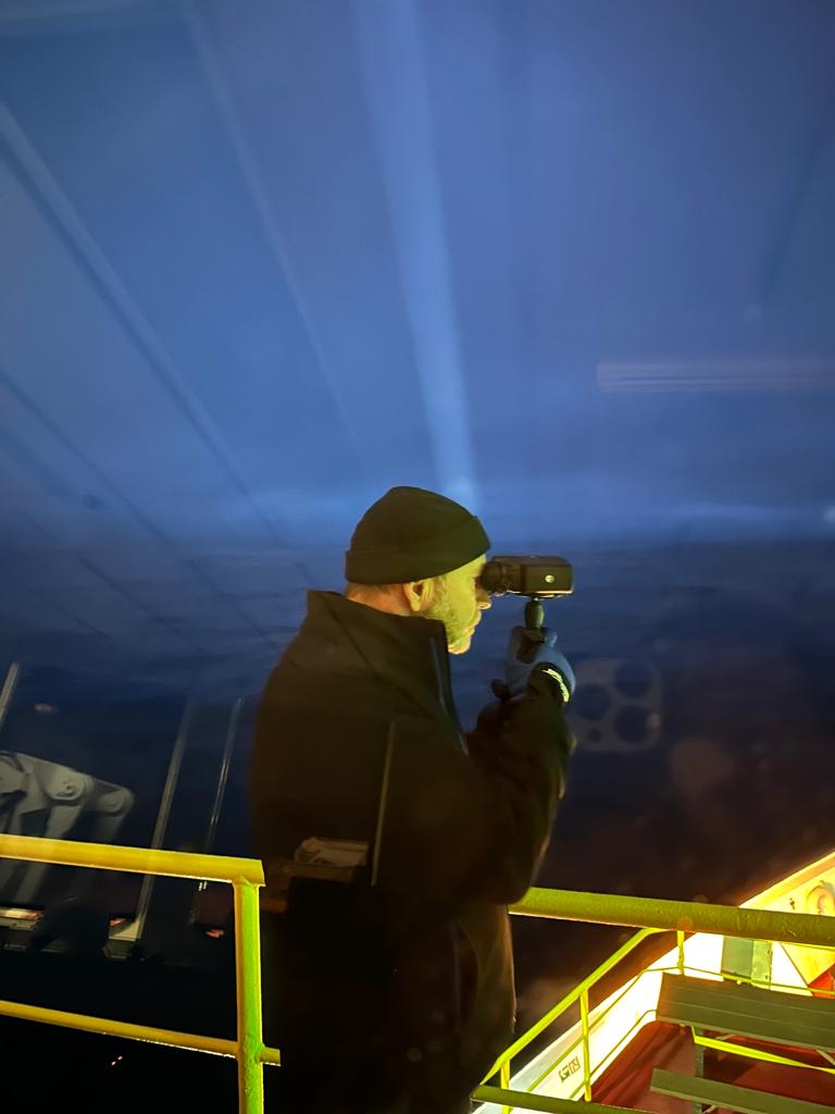 Man standing on a ship deck wearing in black cap and winter coat looking through binoculars.