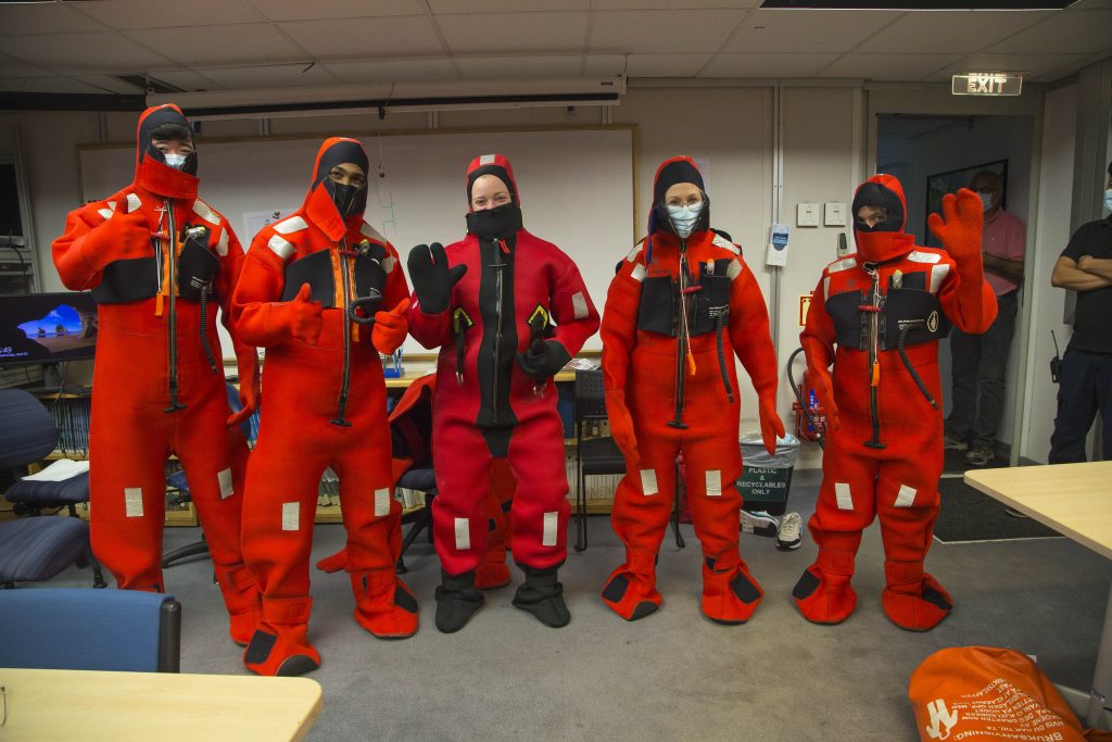 Five people lined up in a row wearing orange survival suits