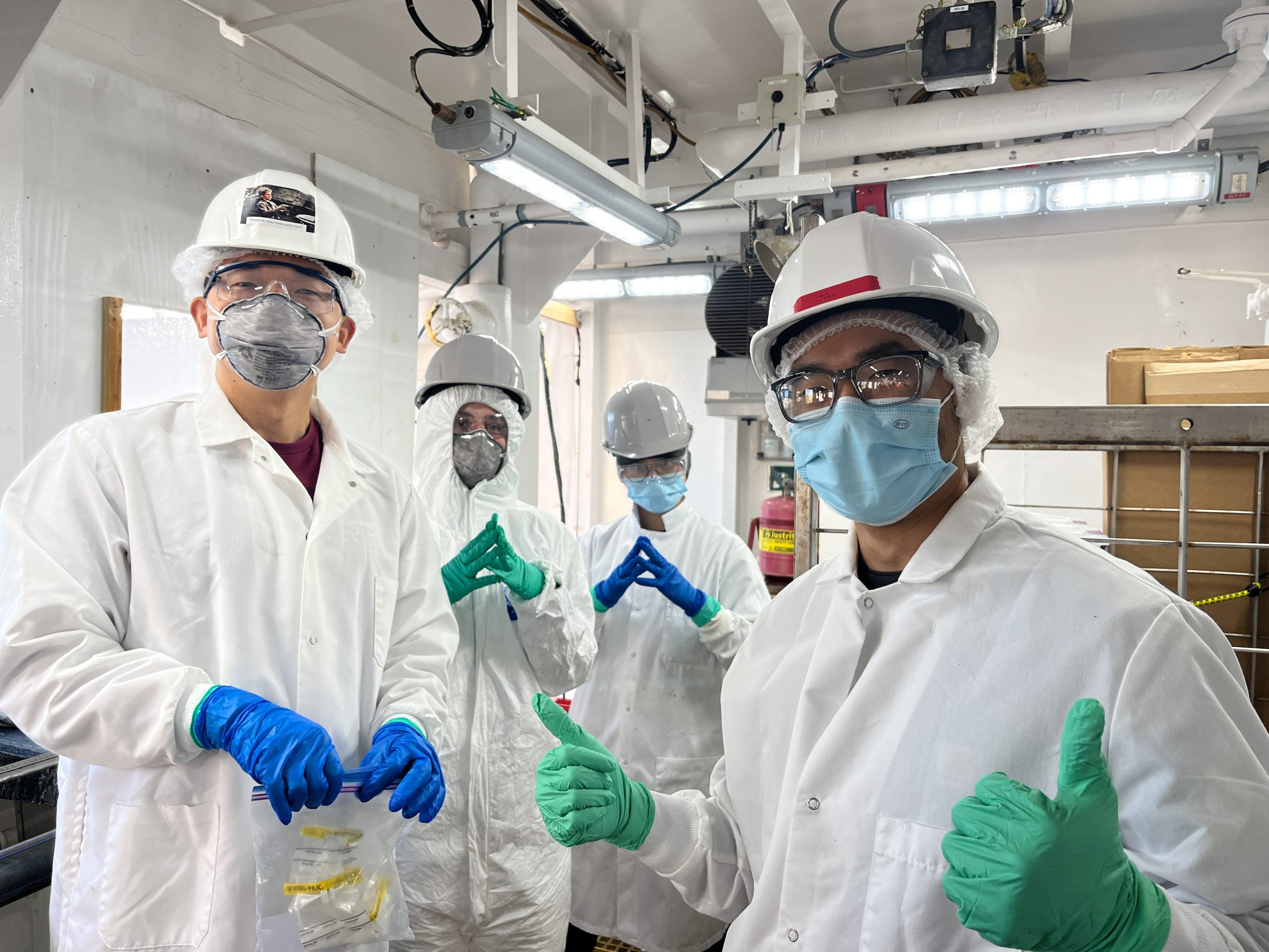 Four persons dressed in white lab coats, hard hats, safety glasses, gloves, and masks