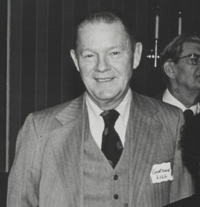 Black and white photo of a smiling man in a three piece striped suit and dark tie and white shirt. He has a receding hairline and a name tag near his left lapel that says "Gordon Lill."