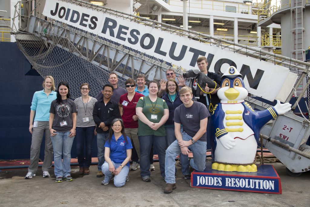 Group of people with a penguin statue posing in front of a ship gangway