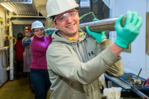 a line of people carrying a long core on their shoulders while wearing hardhats and safety glasses