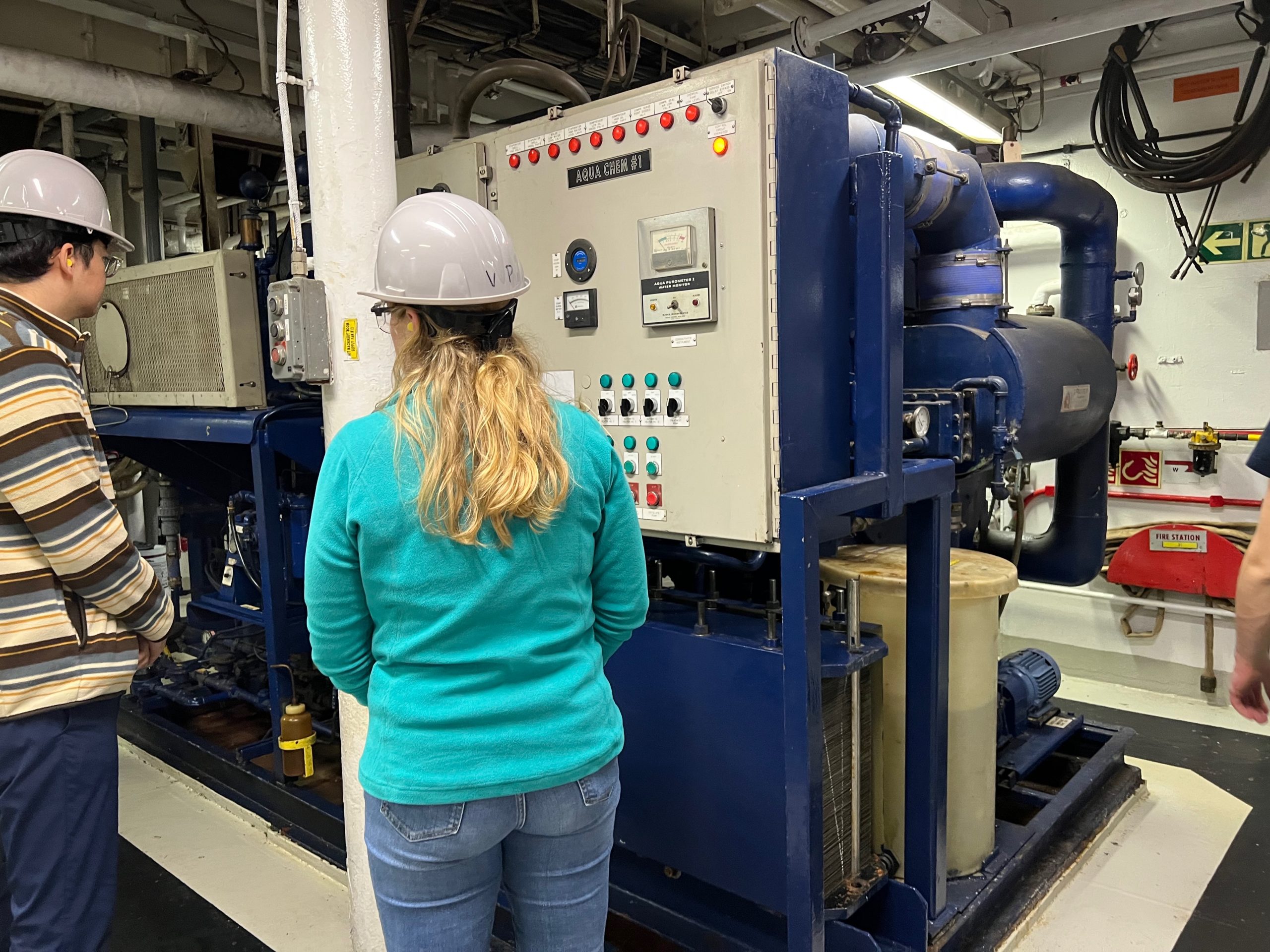 The backs of two persons in hardhats, standing in front of a large navy blue boxy piece of equipment. The equipment has a panel with several buttons. The person on the left is a man with a striped sweater and dark hair. On the right is the woman with long blonde hair and an aqua colored sweater.