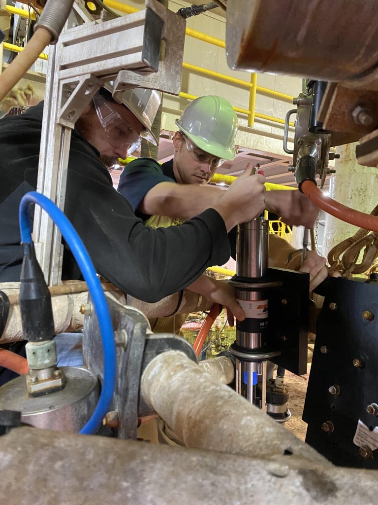 Two people working on a metal cylinder connecting multiple tubes to it and fixing it in place