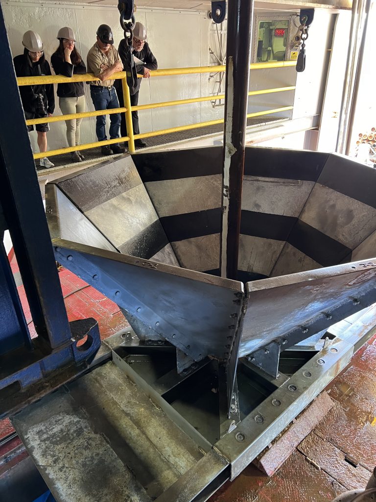 Four people looking over a railing to a large metal funnel sitting on a deck below them