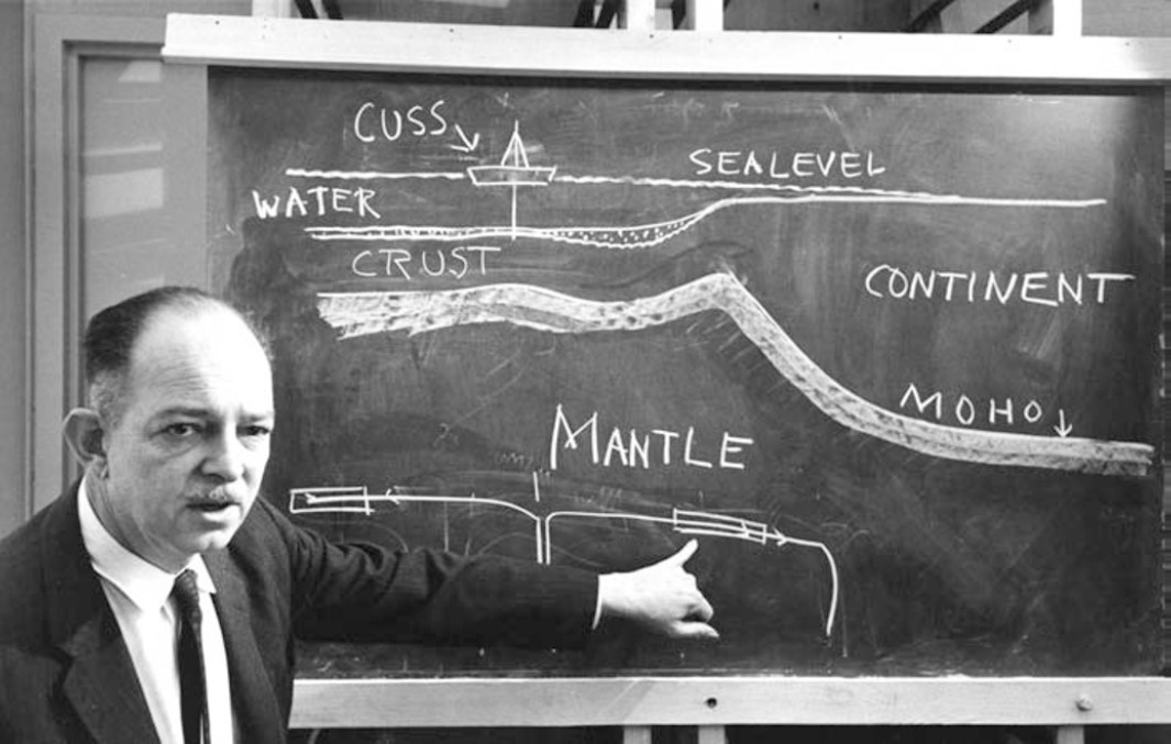 Man pointing to a black board showing a diagram of earth layers and the ocean above.