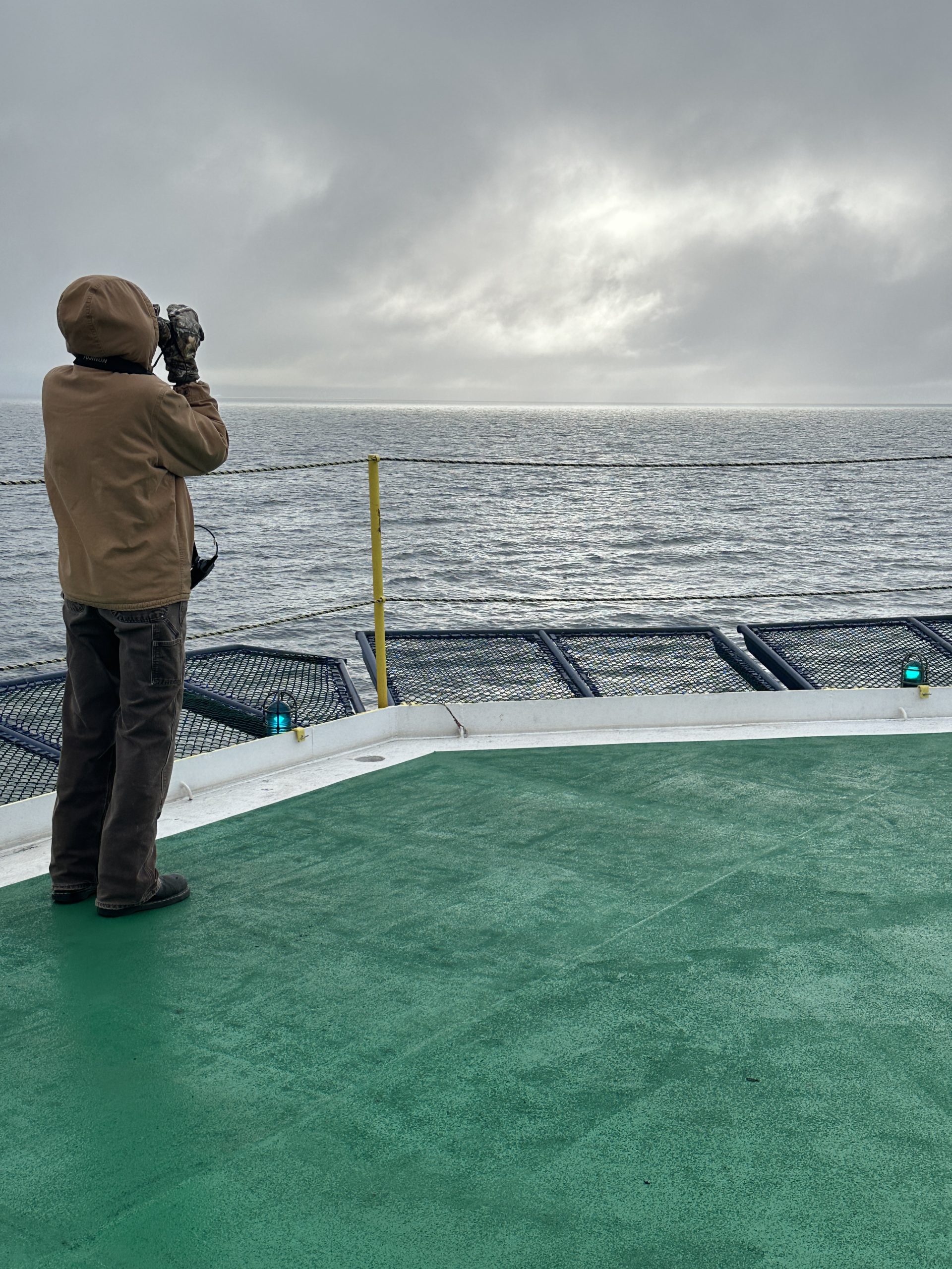 A person stands on the ack of a ship. in a hooded brown coat and is scanning the seas.