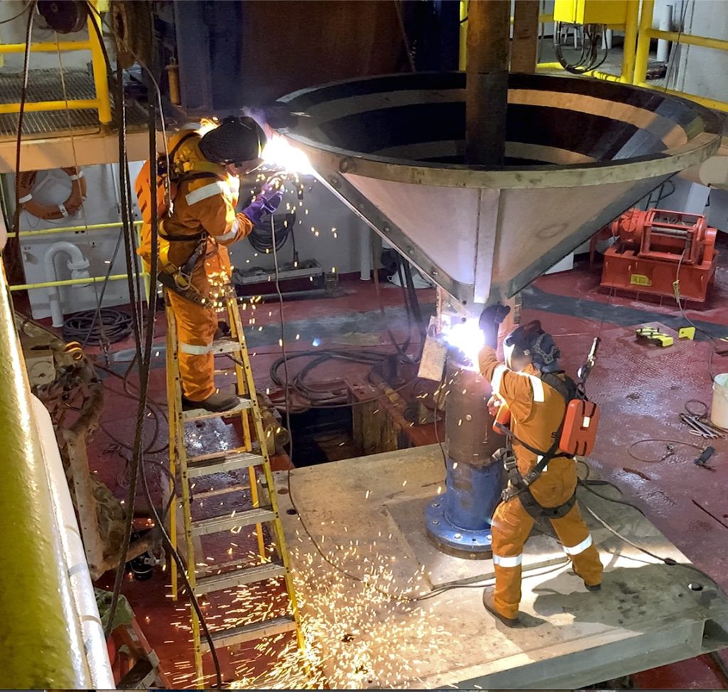 two men in orange jumpsuits working on a large metal funnel, one man welding the top part of the funnel while the other is welding the bottom part