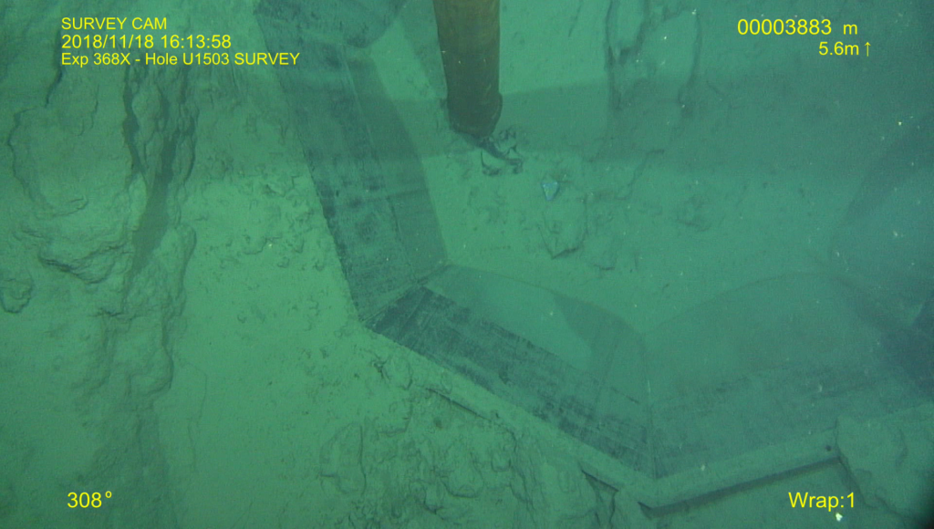 Underwater image of the seafloor with a partially sunken funnel covered in seafloor sediment. Written on the image on top left: Survey Cam, 2018/11/18 16:13:58, Exp 368X- Hole U1503 Survey