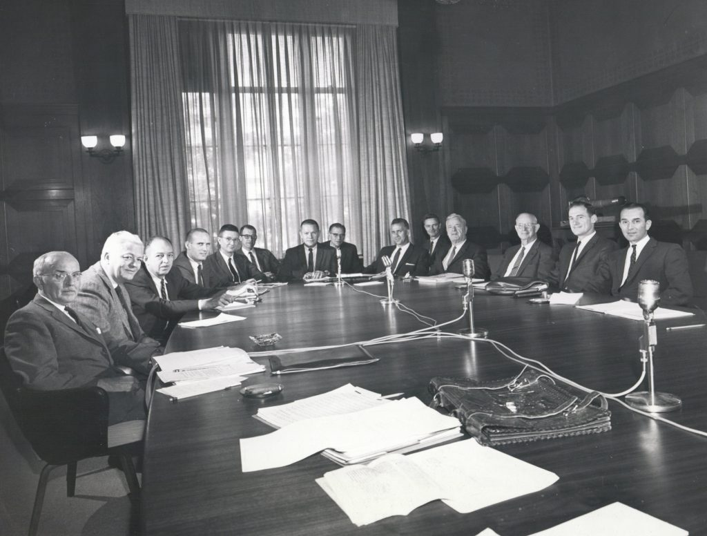 Fourteen men in suits and ties sitting around an oval table looking at the camera