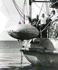 Black and white photo of approximately four men standing at the bow of ship behind a hoisted large lenticular buoy.