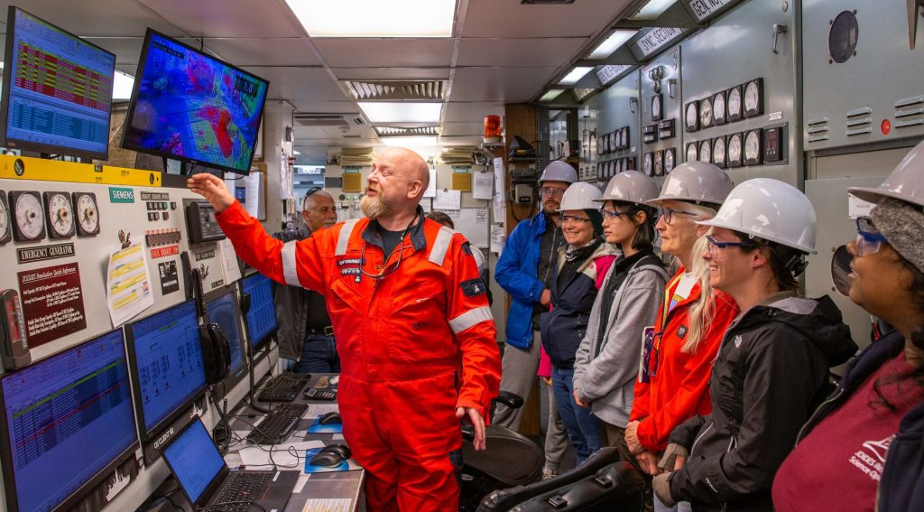 In a ship's engine room that is lined with dials and computers, a smiling bald man with a blonde-gray beard, wearing an orange utility suit is gesturing towards a computer screen showing a chart with red, yellow and green rows and printed text, Several persons in hardhats are looking on.A Siem crewmember shows the engine room, including the water purification controls to a group of scientists. (Credit: Justin Dodd, IODP JRSO, MerlinOne photo archive, CC BY 4.0)