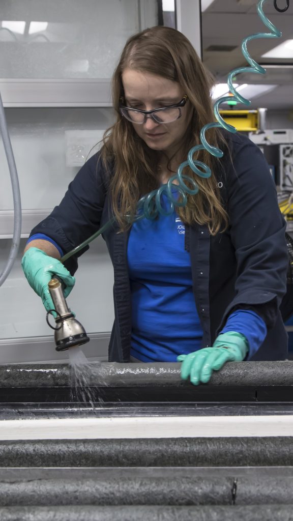 Woman with long brown hair parted on the side is wearing a bright blue long sleeved shirt and an unbuttoned open navy shirt, and light green rubber gloves. She is holding a water nozzle that she is using to spray a core of dark gray rock.