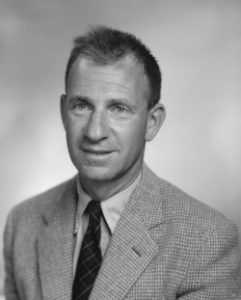 Black and white photograph of a man with crewcut and checked sport coat, light colored button down shirt and dark tie with a checked pattern.