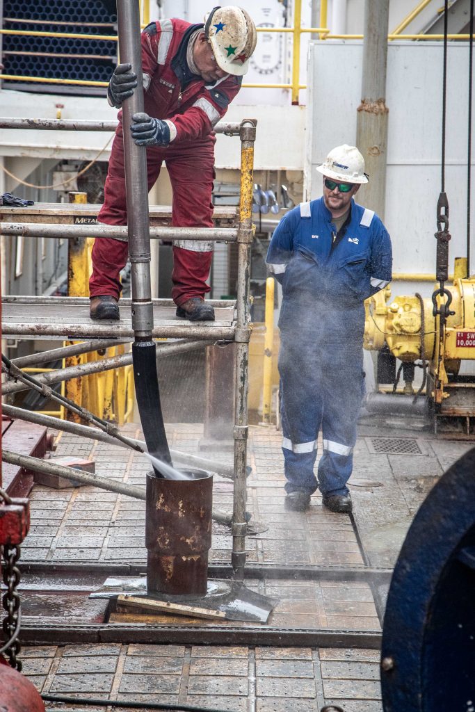 Two men in coveralls on the deck of a ship washing down equipment