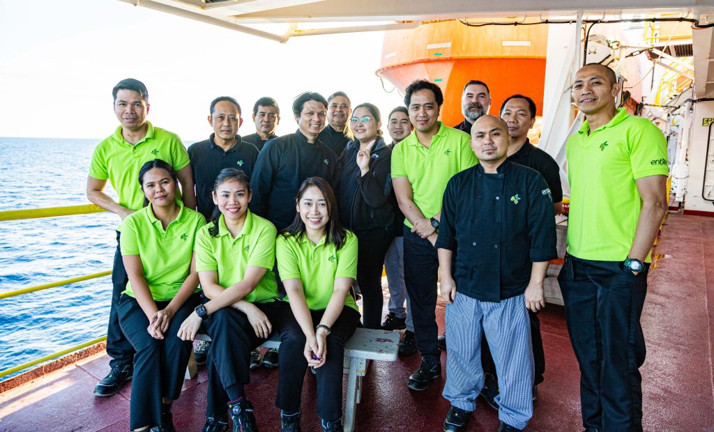 A group of people standing and sitting while posed on the deck of a ship in the ocean.