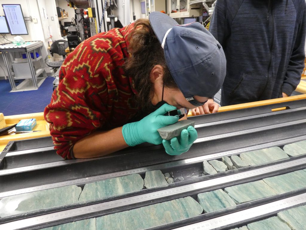 Person using a small hand lens wearing gloves and holding a piece of green rock, leaning over a split core of green rock