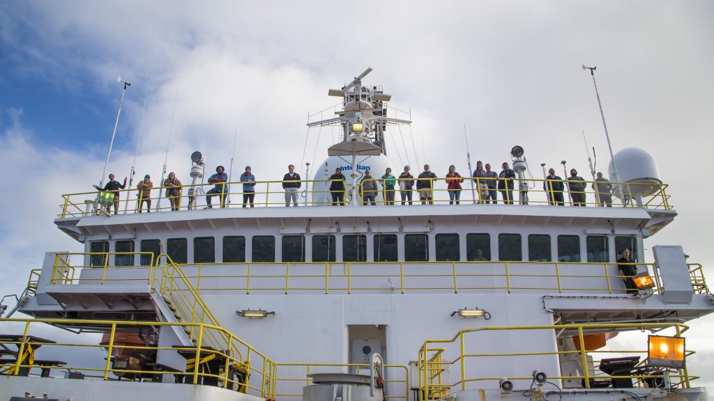 A photo facing the bridge of a ship with people standing on the level above the bridge