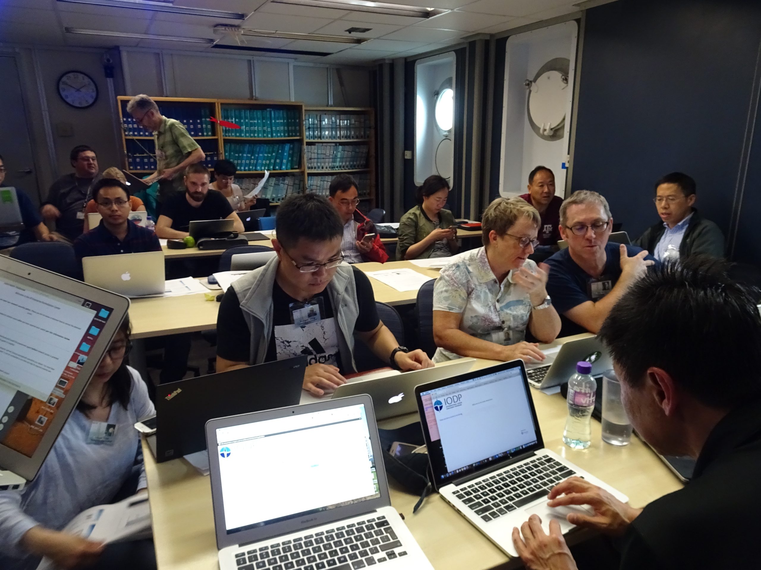 A crowded room filled with people sitting on every side of a table on their laptops.