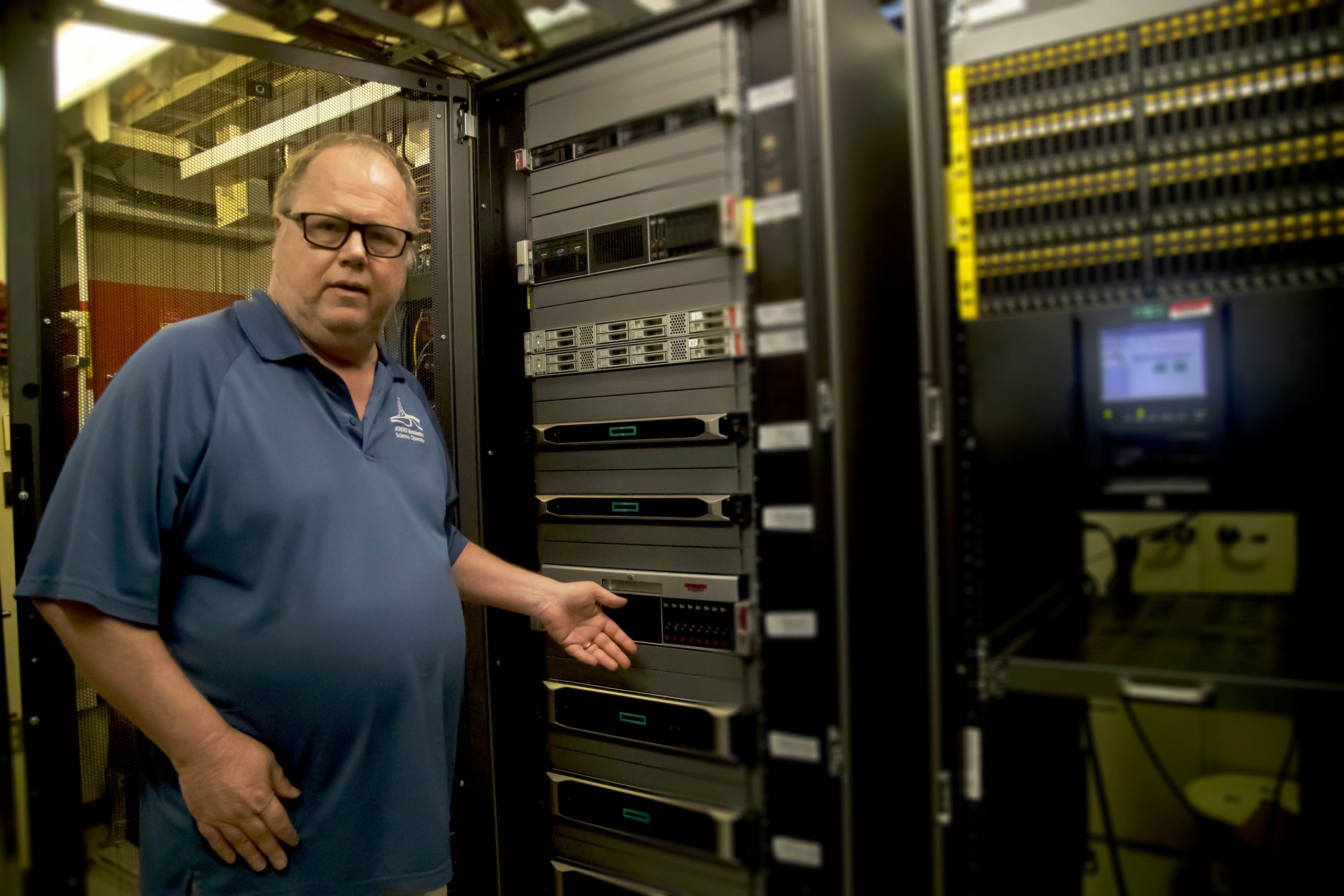 man standing in the computer server room pointing to one of the servers.