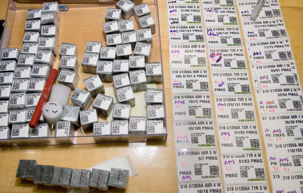 About 75 small boxes with numerical and QR codes sit in the upper right. In the lower left part of the image are six gray rock cube samples with red hatched arrows on them. The right side of the image shows three columns of labels containing numerical codes, expedition information and QR codes.