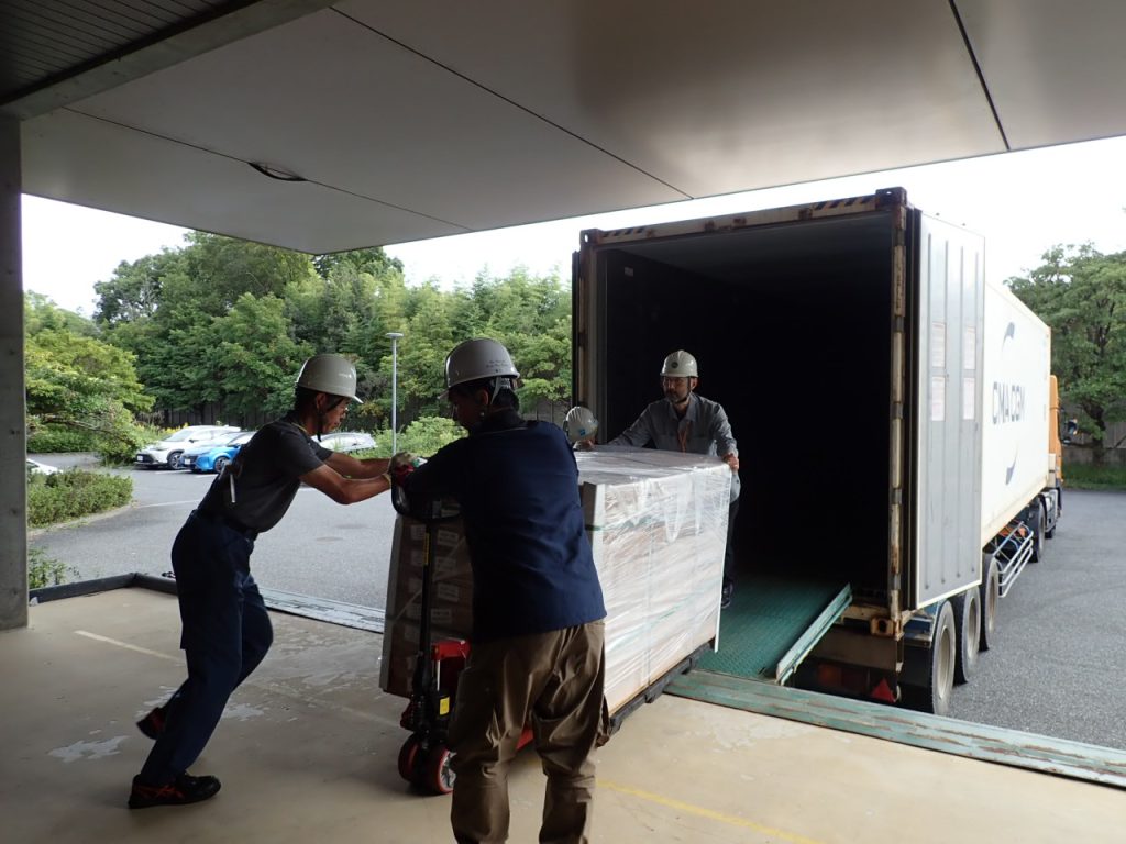 Three people standing in front of a truck moving a big shipment of boxes.