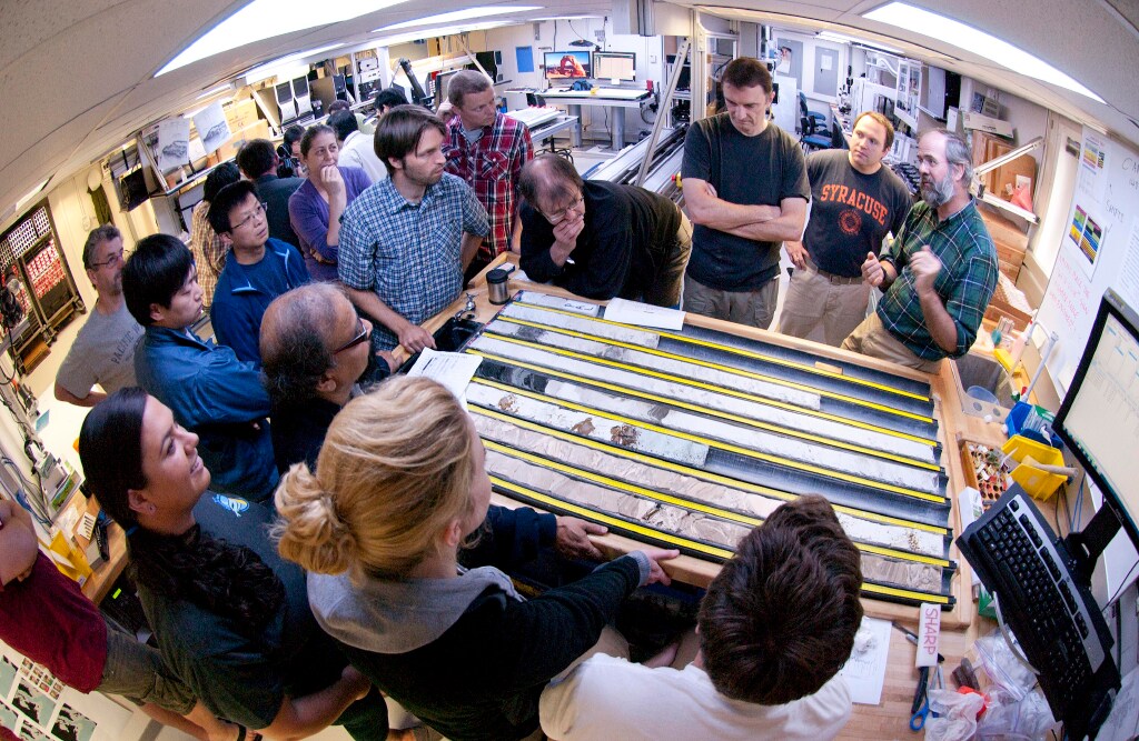 A fish-eye lens view of fourteen scientists looking at and discussing several (seven) cores. They are looking at one person who is gesturing with his hands as if he is making a point.