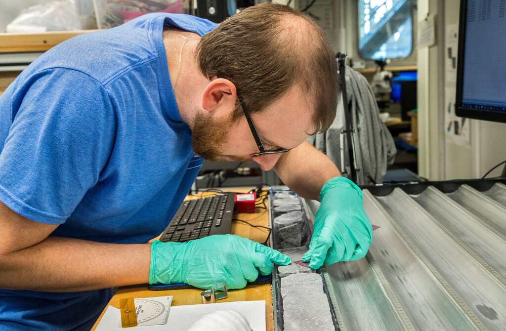 A man with brown hair and glasses is leaning over a rocks in a core. He has on green rubber gloves and is holding a ruler onto the rock.