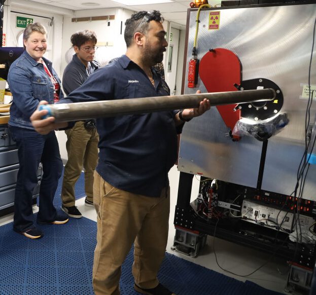 A man with dark hair and a beard places a long core segment into a hole with a red door that slides shut on it. One smiling person looks on and another person is looking at the side (not seen in this image) of this x ray machine.
