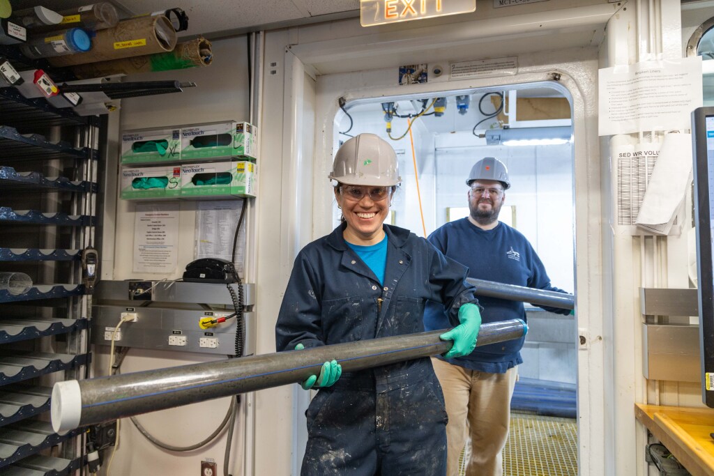 Smiling woman and behind here a smiling man, both wearing hard hats and safety glasses carry core sections from the catwalk into the core lab.